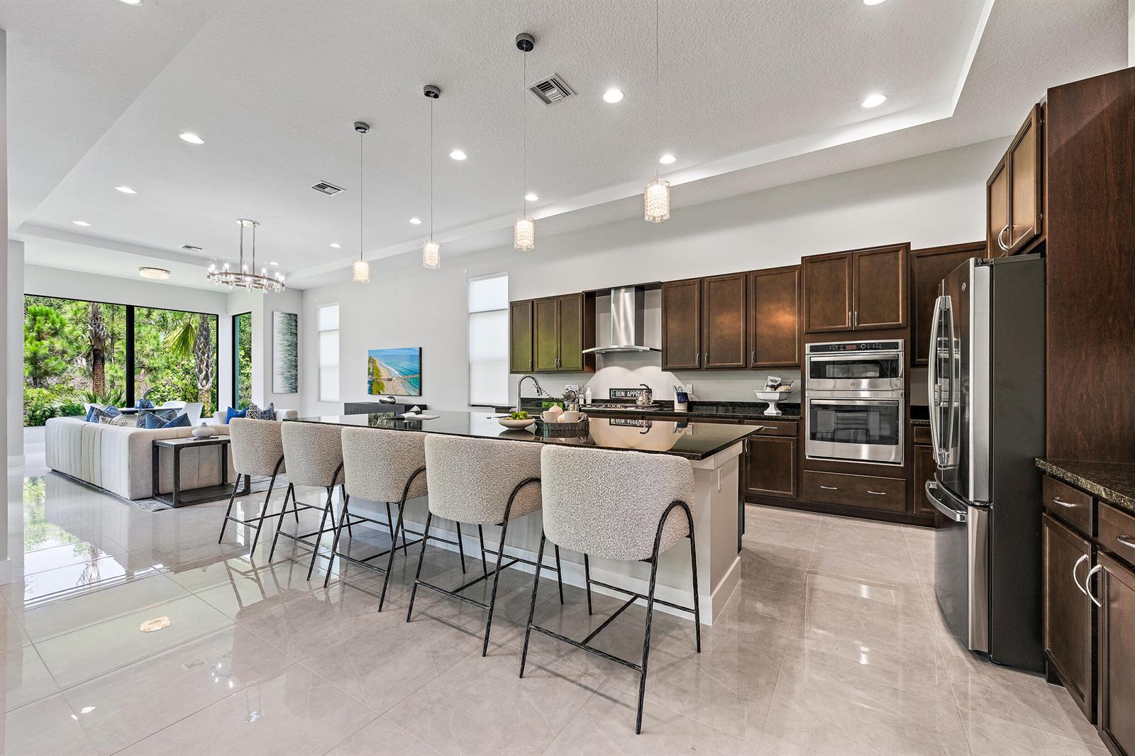 A modern kitchen with dark wood cabinets, a large island, and bar seating overlooks a living room — 13358 Artisan Circle, Palm Beach Gardens FL 33418, PALM BEACH GARDENS, FL