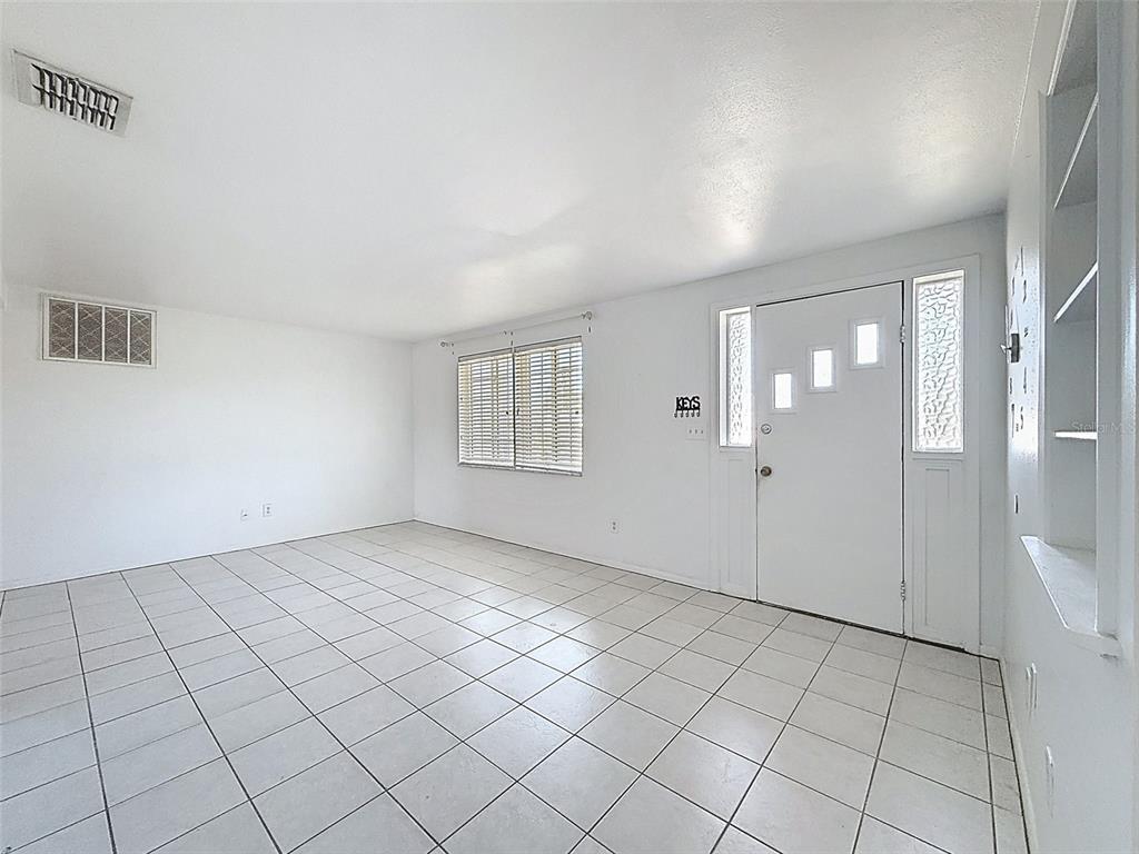 A bright, empty room features white walls, tiled floors, a window with blinds, and an entryway — 4130 CRESTFIELD AVENUE, HOLIDAY, FL