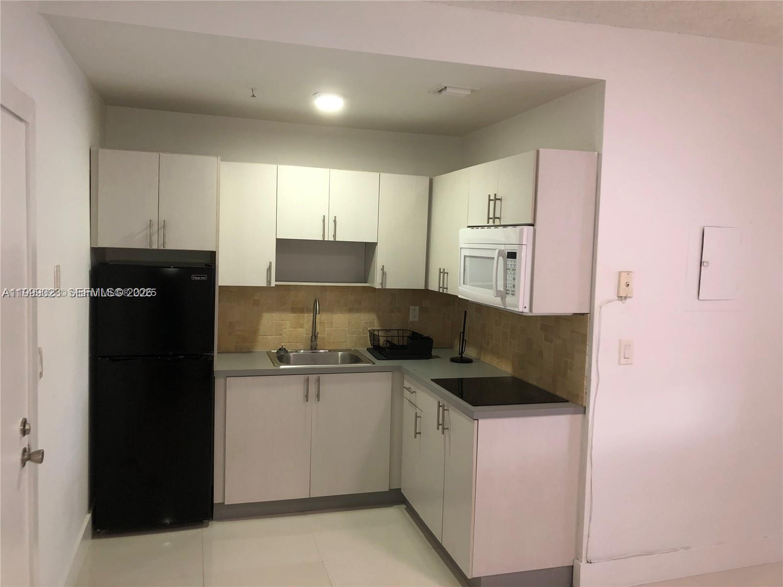 A small kitchen features white cabinets, a black refrigerator, a stainless steel sink, and tile backsplash — 2730 Collins Ave # 310, Miami Beach FL 33140, MIAMI BEACH, FL