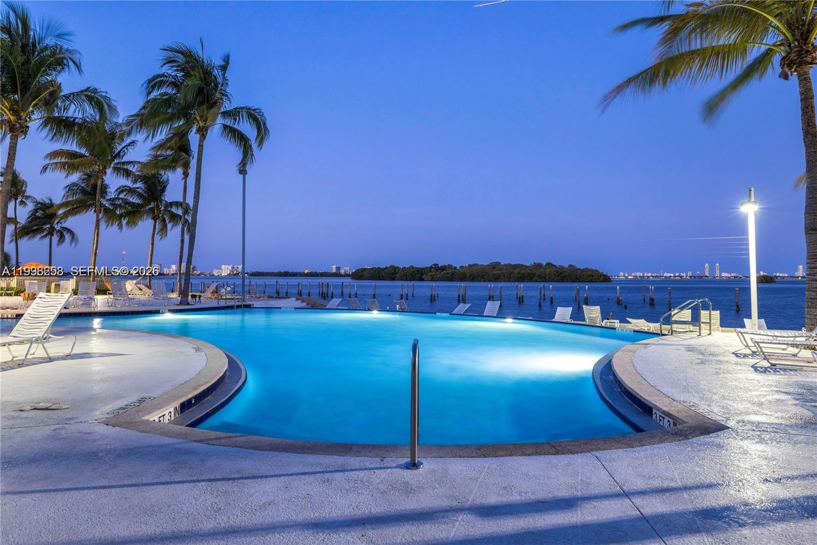An illuminated swimming pool overlooks a bay with palm trees and a distant city skyline — 680 NE 64th St # A300, Miami FL 33138, MIAMI, FL
