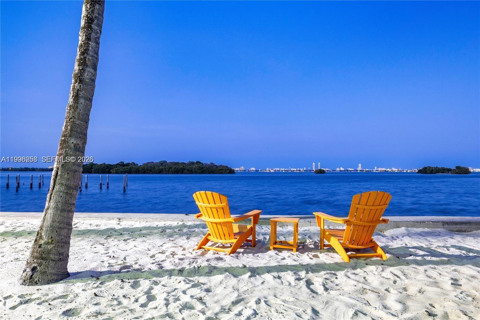 Two yellow chairs and a small table sit on a sandy beach overlooking water — 680 NE 64th St # A300, Miami FL 33138, MIAMI, FL