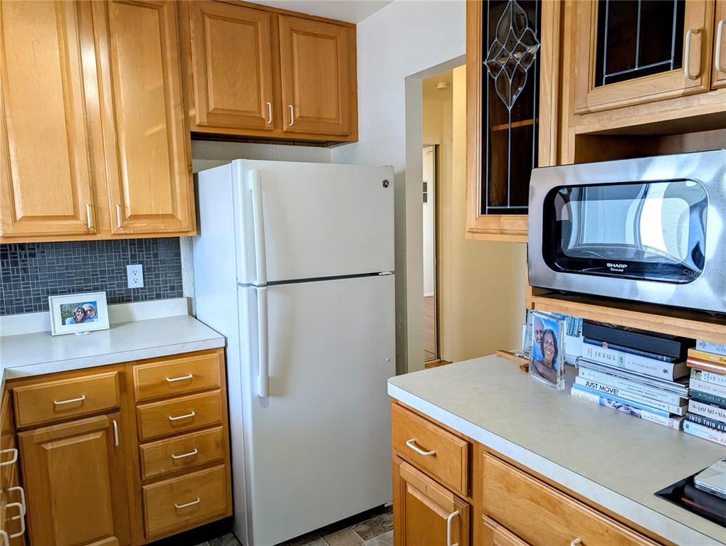 A kitchen features wooden cabinets, a white refrigerator, a microwave, and a tiled backsplash — 1545 PLEASANT ROAD UNIT H-23, BRADENTON, FL