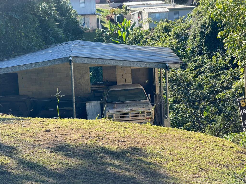 A weathered pickup truck is parked under a simple metal-roofed carport in a grassy area — CAFARNAUM SOLAR 118, AIBONITO, PR