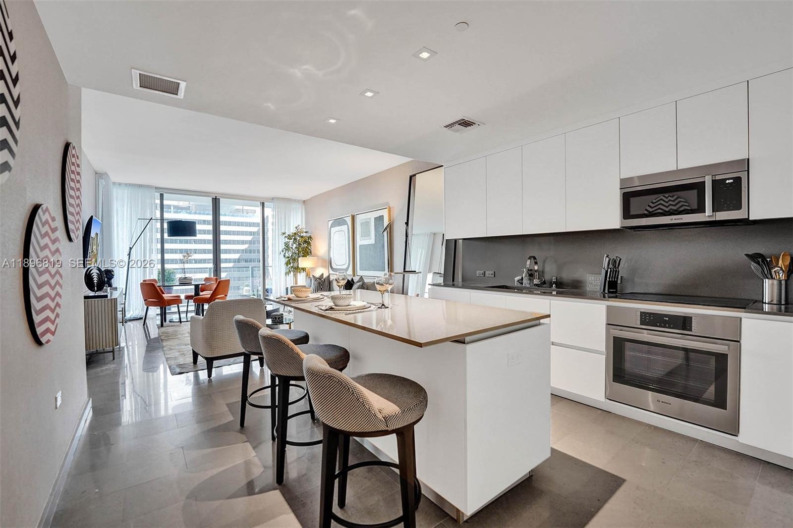 A modern kitchen with white cabinetry and a central island overlooks a bright living area — 88 SW 7th St # 1408, Miami FL 33130, MIAMI, FL