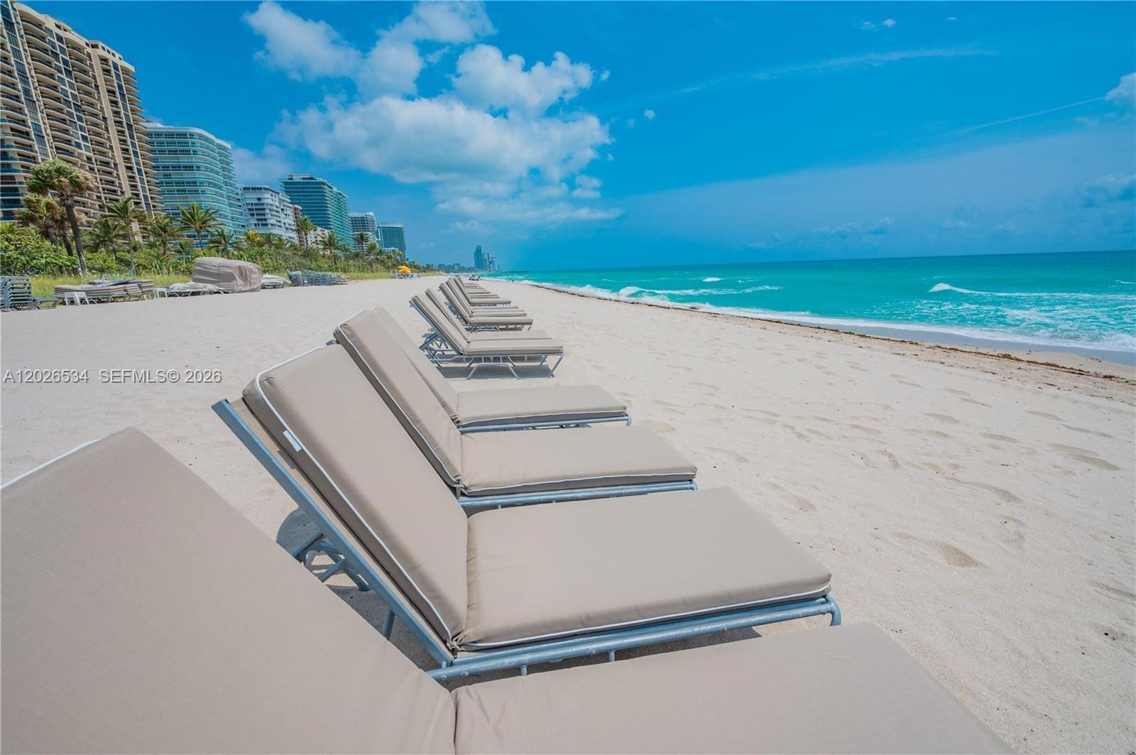 A row of tan lounge chairs sits on a sandy beach near high-rise buildings — 9801 Collins Ave # 6P, Bal Harbour FL 33154, BAL HARBOUR, FL