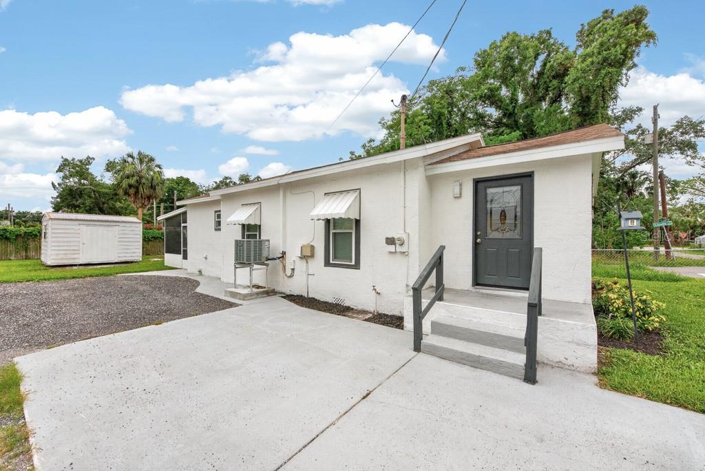 A white stucco house exterior features a concrete patio, a gray front door, and a shed — 6101 ANNA AVENUE, GIBSONTON, FL