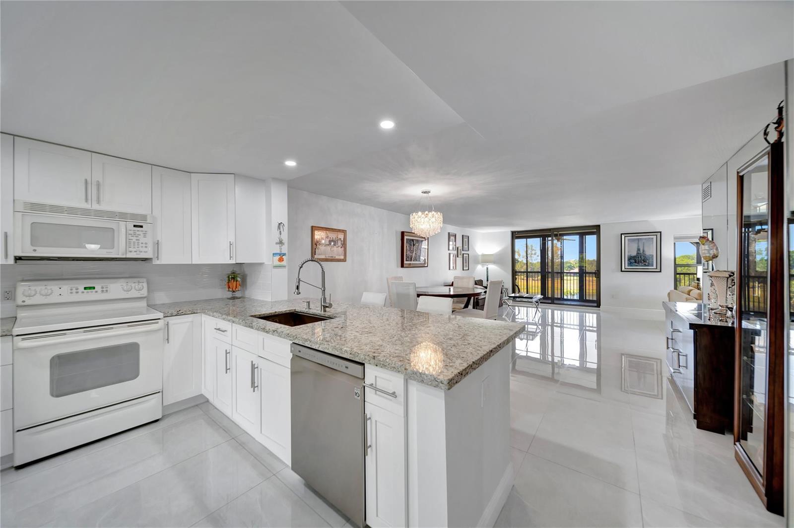 A modern white kitchen with granite countertops opens into a dining area with balcony views — 3521 Environ Boulevard # B410, Lauderhill FL 33319, LAUDERHILL, FL