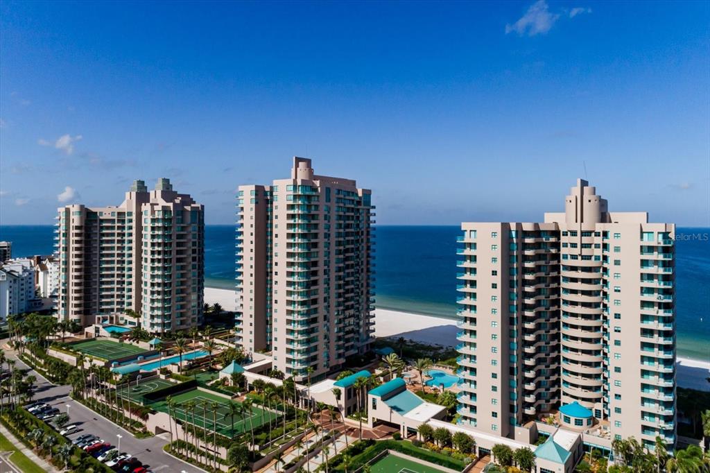 An aerial view shows three tall beachfront residential towers with swimming pools and tennis courts — 1520 GULF BOULEVARD UNIT 1404, CLEARWATER BEACH, FL