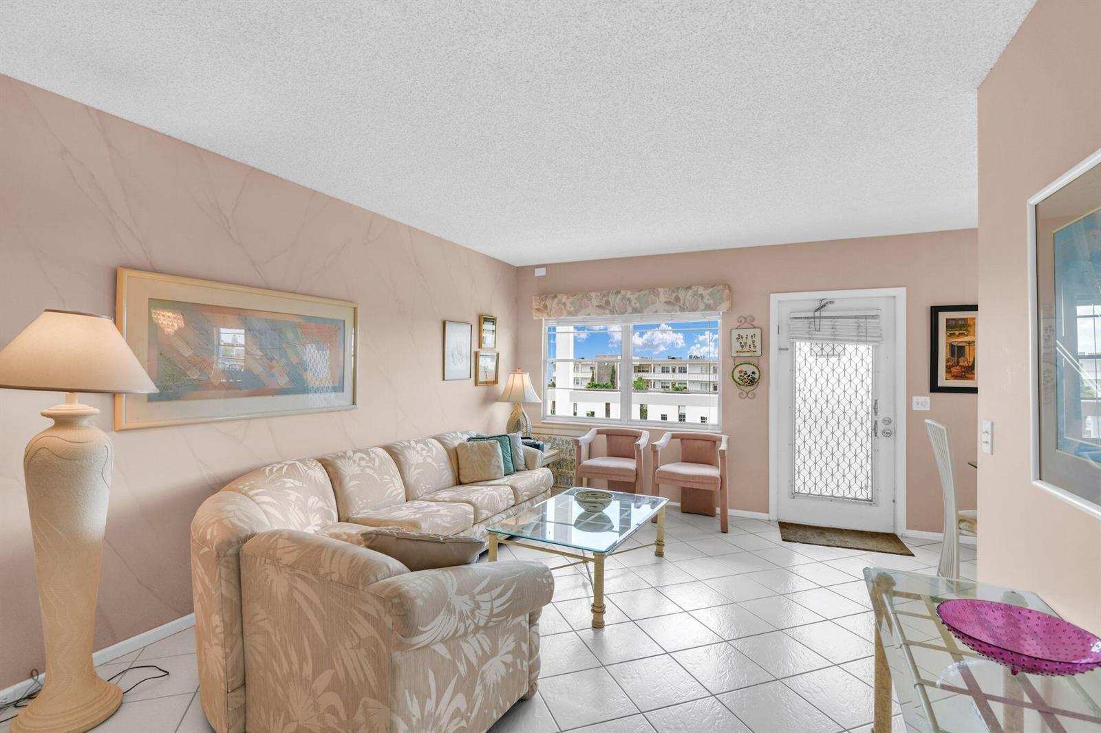 A living room with tile floors, a floral sofa, glass coffee table, and a front door — 4053 Wolverton C, Boca Raton FL 33434, BOCA RATON, FL
