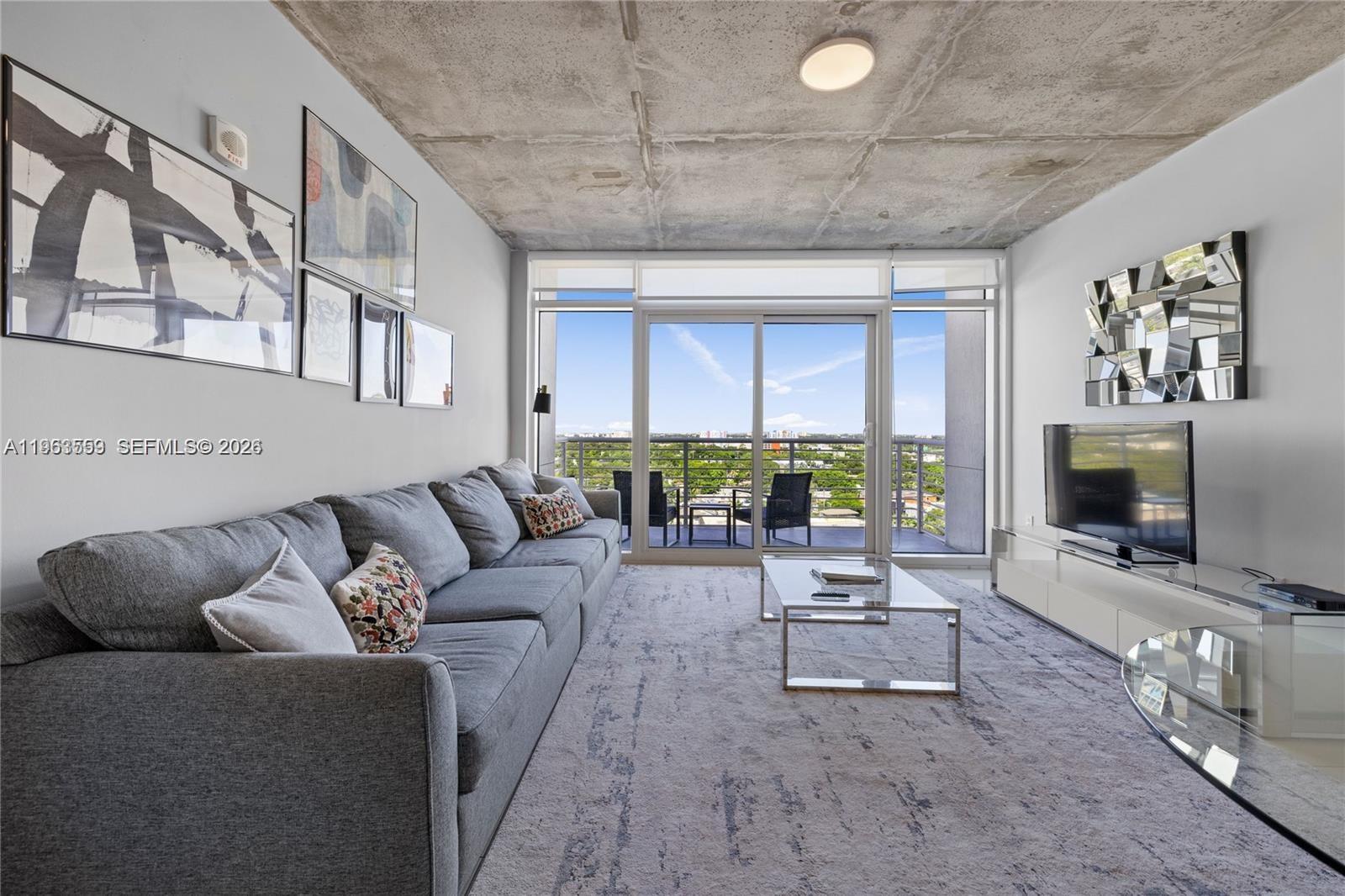 A modern living room features a gray sofa, concrete ceiling, and a balcony with views — 3250 NE 1st Ave # 1113, Miami FL 33137, MIAMI, FL