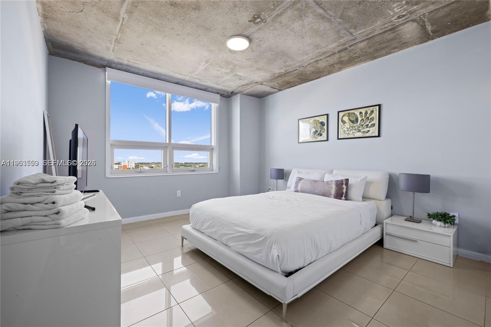 A bedroom features a white bed, a concrete ceiling, and a large window with a view — 3250 NE 1st Ave # 1113, Miami FL 33137, MIAMI, FL