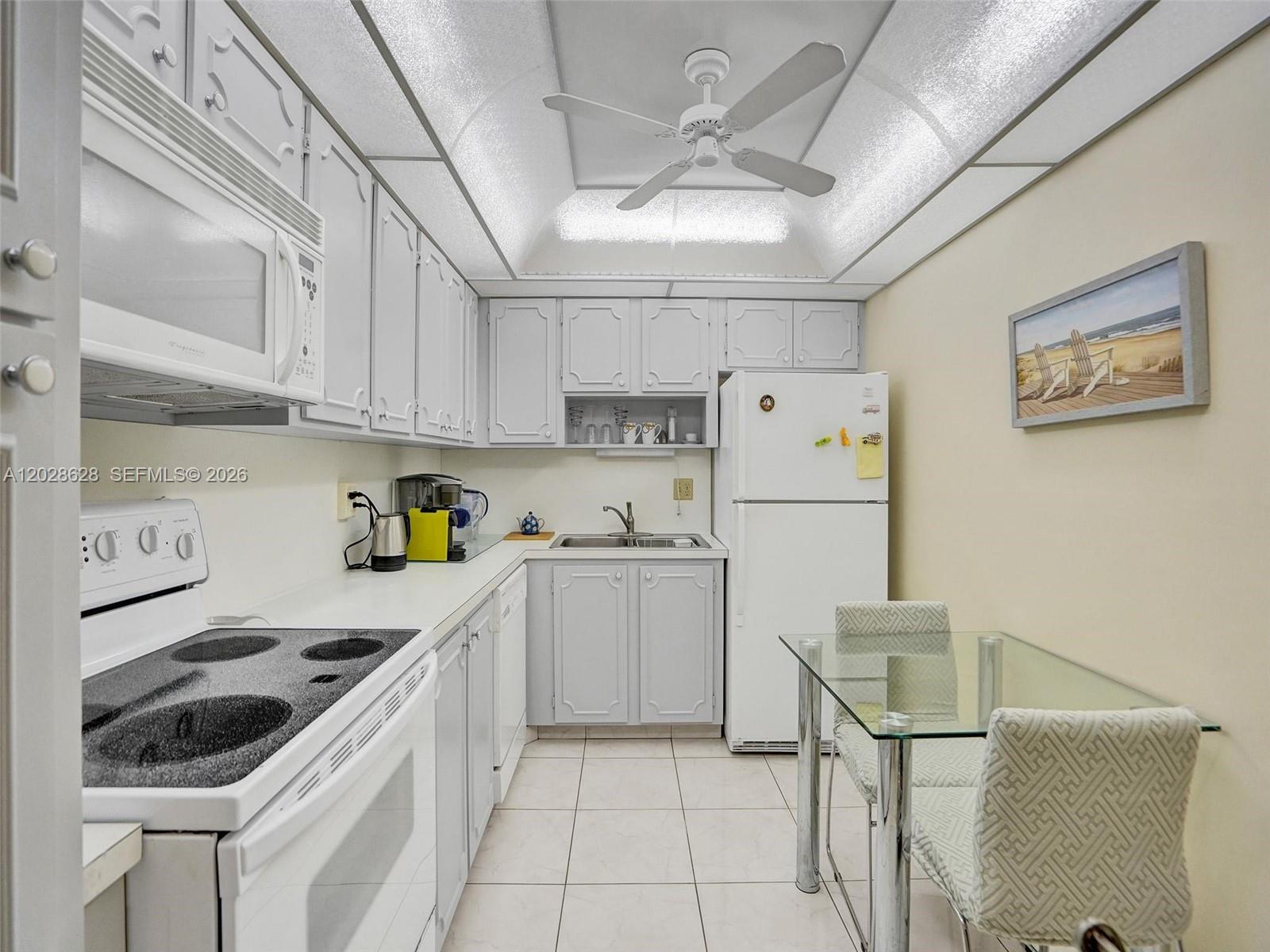 A small kitchen features white cabinets, white appliances, tile flooring, and a glass dining table — 3113 S Ocean Dr # 603, Hallandale Beach FL 33009, HALLANDALE BEACH, FL