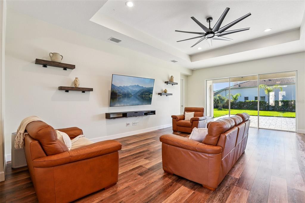 A living room features leather furniture, a wall-mounted television, floating shelves, and wood-look flooring — 3326 ACACIA BAY AVENUE, WESLEY CHAPEL, FL