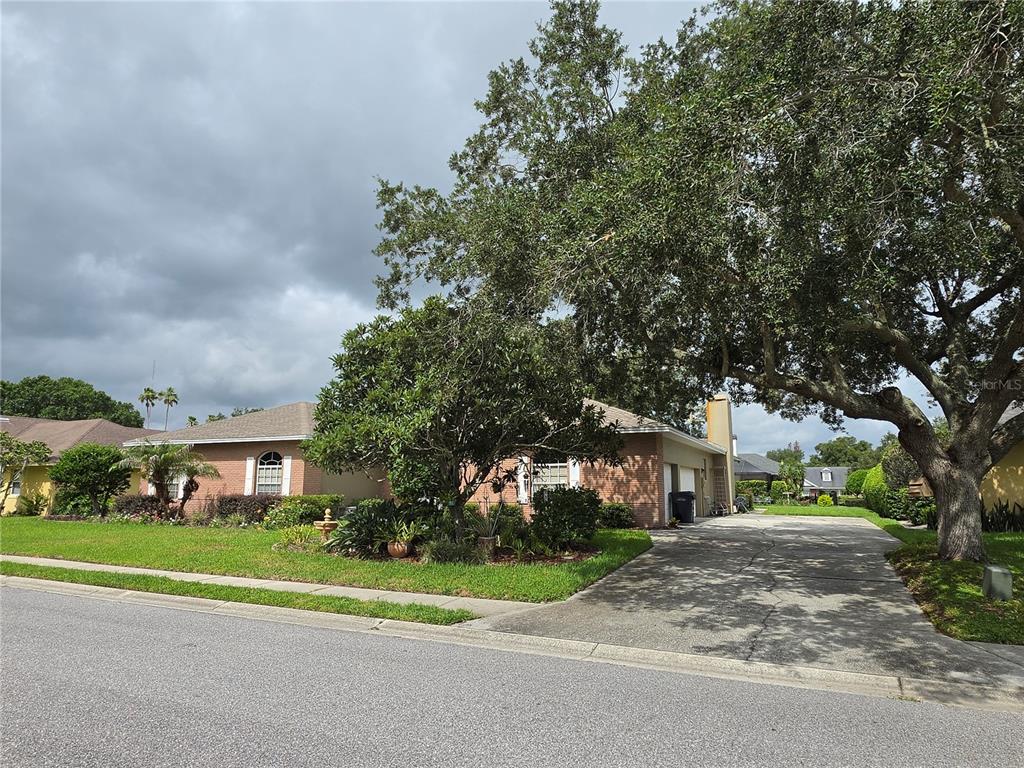 A single-story brick house with a concrete driveway and large trees under a cloudy sky — 6523 CREWS LAKE HILLS LOOP E, LAKELAND, FL