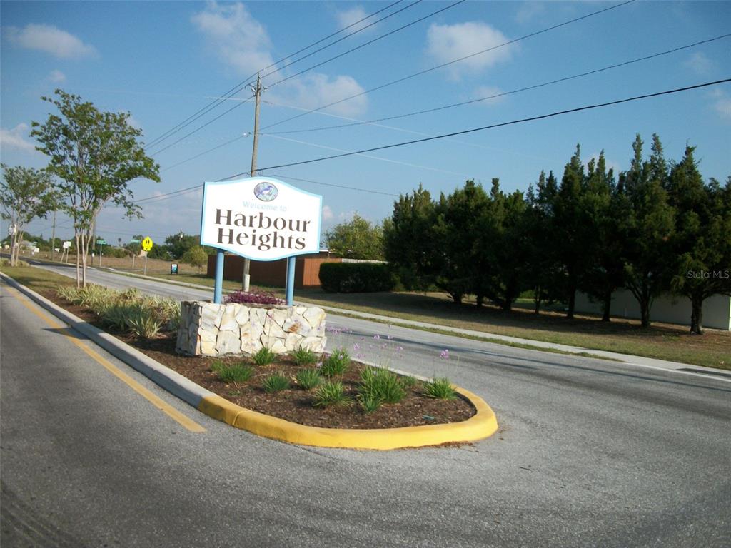 A stone monument sign welcomes visitors to the Harbour Heights community along a paved road — 1228 HARBOUR HIBISCUS COURT, PUNTA GORDA, FL