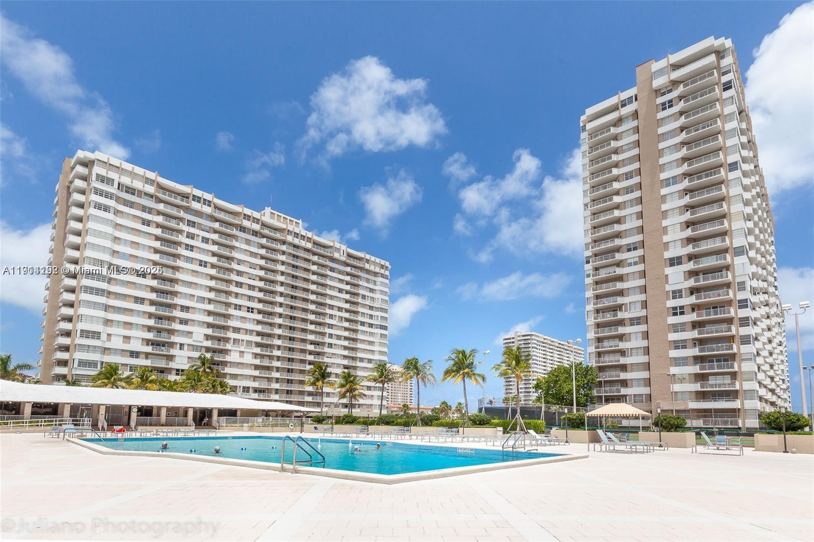 A swimming pool sits on a patio between two tall residential apartment buildings — 1965 S Ocean Dr # M, Hallandale Beach FL 33009, HALLANDALE BEACH, FL