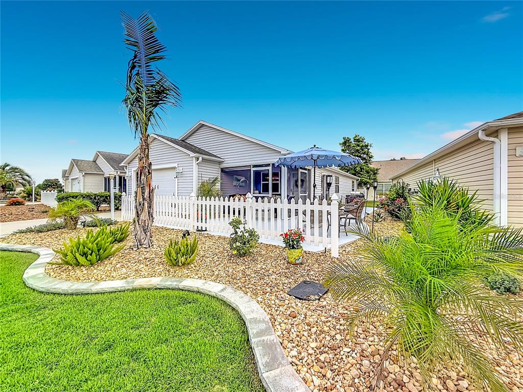 A house exterior features a white picket fence, a patio with an umbrella, and landscaping — 1648 ABASCO COURT, THE VILLAGES, FL