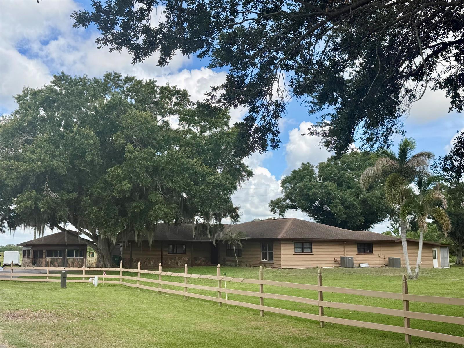 A single-story house with brown siding and a large oak tree sits behind a fence — 1631 SE 24th Boulevard, Okeechobee FL 34974, OKEECHOBEE, FL