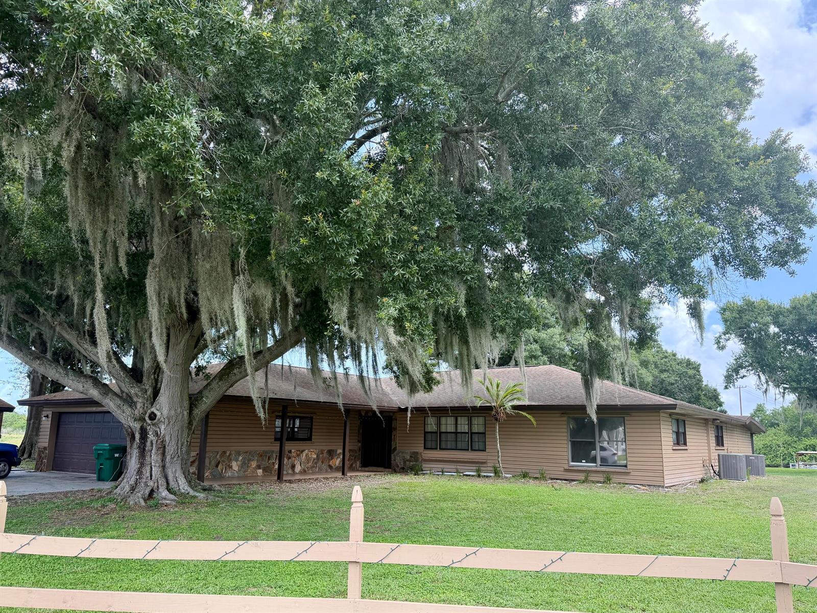 A single-story house with brown siding and stone accents sits behind a large oak tree — 1631 SE 24th Boulevard, Okeechobee FL 34974, OKEECHOBEE, FL