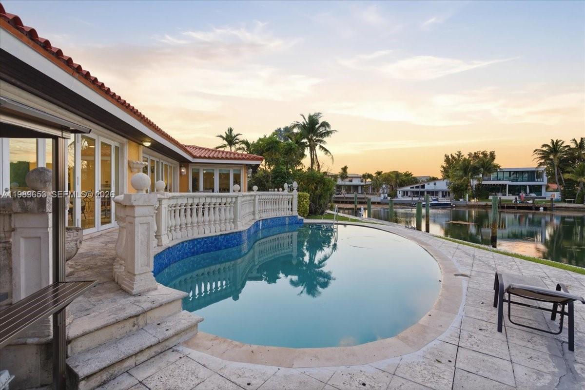 A curved swimming pool sits on a stone patio beside a house with a balustrade — 1249 Biscaya Dr, Surfside FL 33154, SURFSIDE, FL