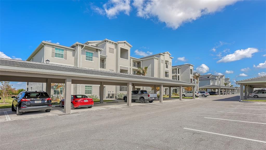 A multi-story apartment building features covered parking spaces in the foreground under a bright sky — 12180 WELLEN GOLF STREET UNIT 202, VENICE, FL