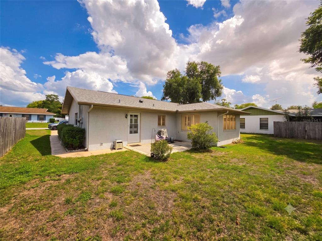 The exterior of a light-colored, single-story house features a small patio and grassy backyard — 7047 WINDMERE ROAD, BROOKSVILLE, FL