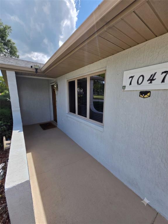 A covered concrete porch leads to a front door and a window on a stucco house — 7047 WINDMERE ROAD, BROOKSVILLE, FL