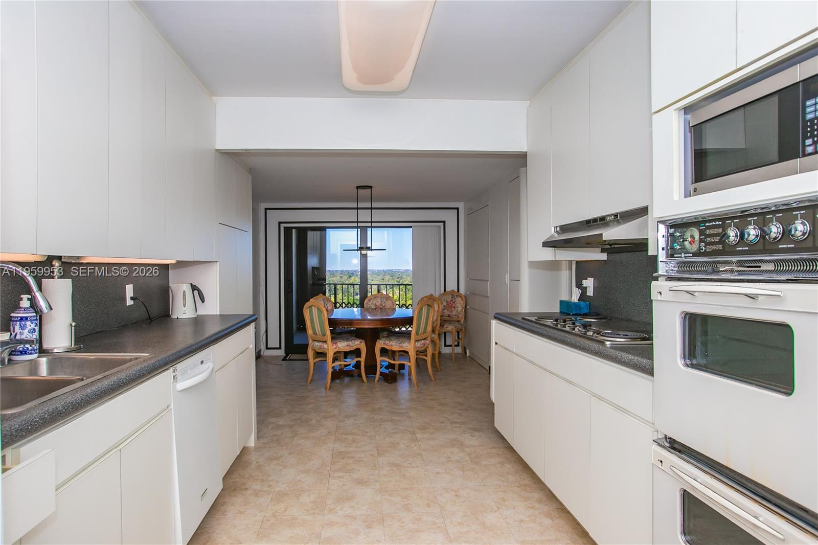 A galley kitchen with white cabinets and dark countertops leads to a dining area — 1800 NE 114th St # 1810, Miami FL 33181, MIAMI, FL