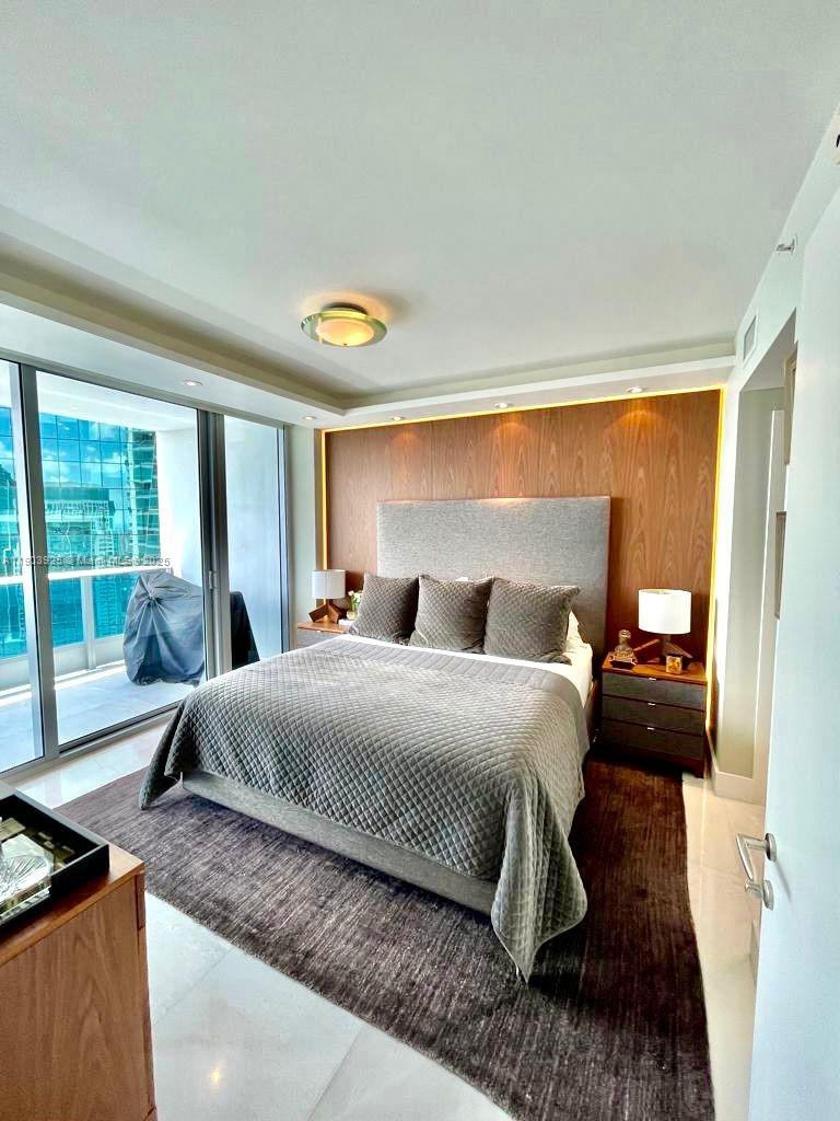 A modern bedroom features a gray upholstered bed, wood-paneled accent wall, and floor-to-ceiling windows — 200 BISCAYNE BOULEVARD WAY # 4310, Miami FL 33131, MIAMI, FL