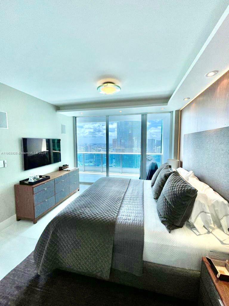 A modern bedroom features a king bed, a wall-mounted television, and floor-to-ceiling windows overlooking city buildings — 200 BISCAYNE BOULEVARD WAY # 4310, Miami FL 33131, MIAMI, FL