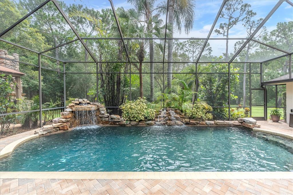 A screened-in swimming pool features a stone waterfall and lush landscaping in a backyard setting — 5328 MACOSO COURT, NEW PORT RICHEY, FL