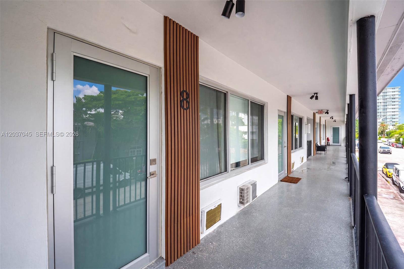 An exterior walkway features a glass door, a wooden slat accent wall, and window-mounted air conditioners — MIAMI BEACH, FL