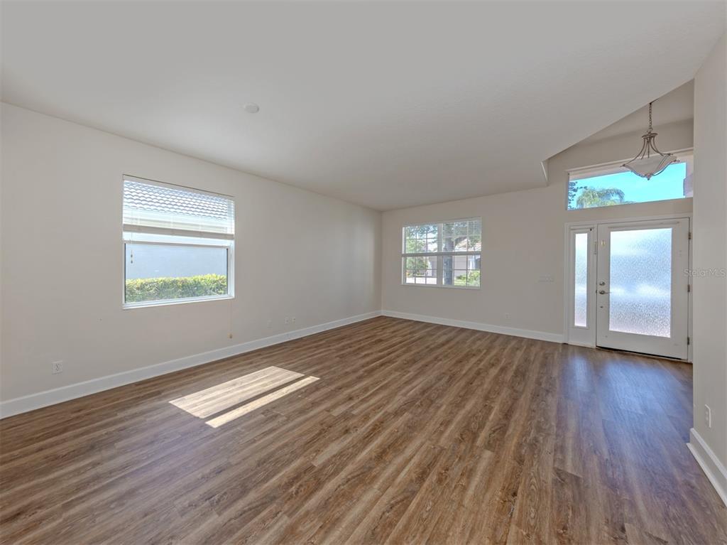 An empty room features light-colored walls, wood-look flooring, multiple windows, and a glass-paneled front door — 11626 DANCING RIVER DRIVE, VENICE, FL