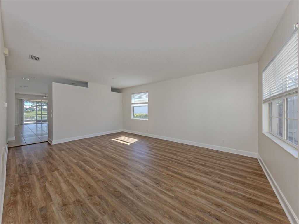 An empty living room features wood-look flooring, neutral walls, and windows with white blinds — 11626 DANCING RIVER DRIVE, VENICE, FL