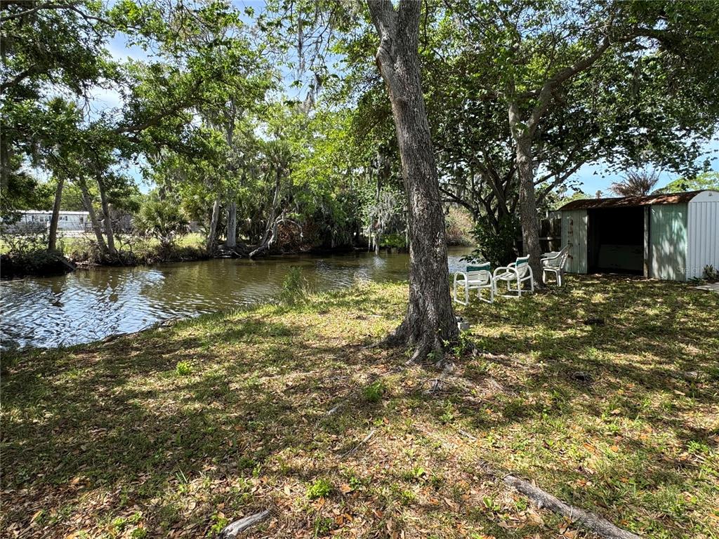 A grassy backyard features a small shed, several chairs, and trees overlooking a canal — 5406 SYDNEY STREET, PORT ORANGE, FL