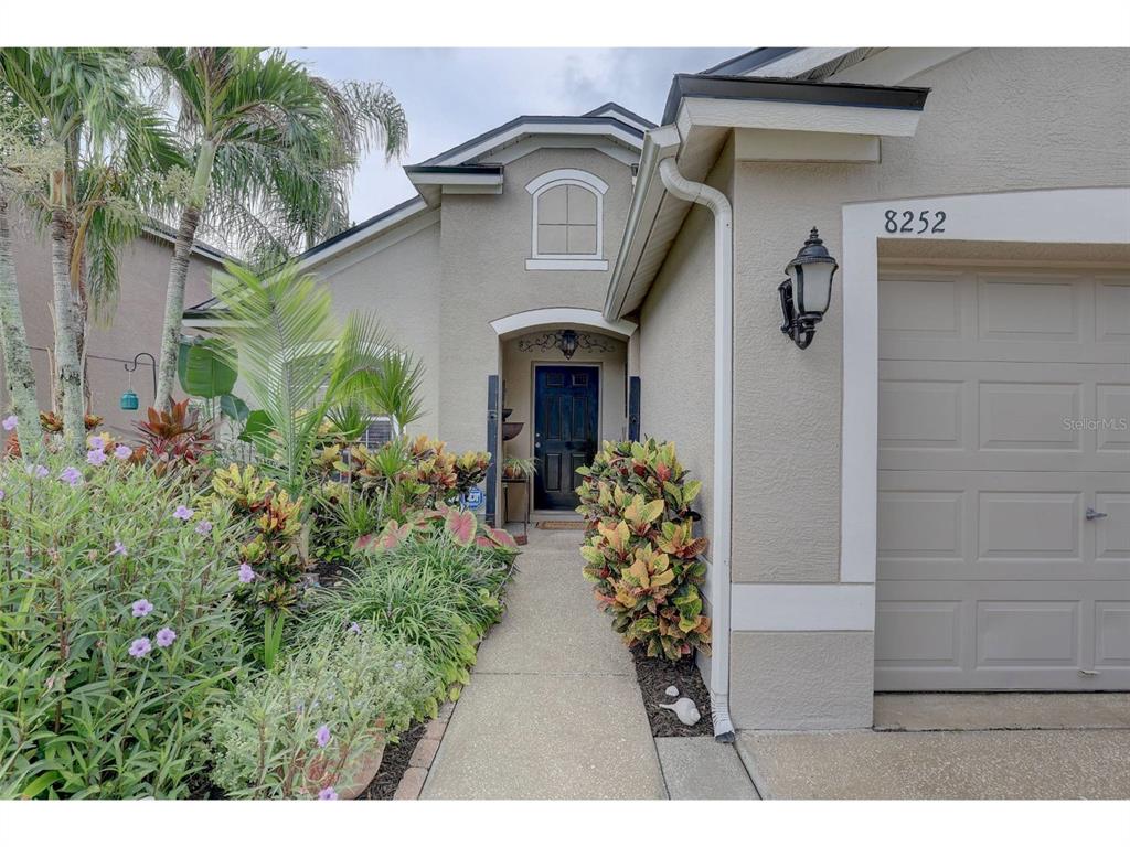 The exterior of a single-story house features a concrete walkway, a garage, and landscaped gardens — 8252 CRESCENT MOON DRIVE, NEW PORT RICHEY, FL