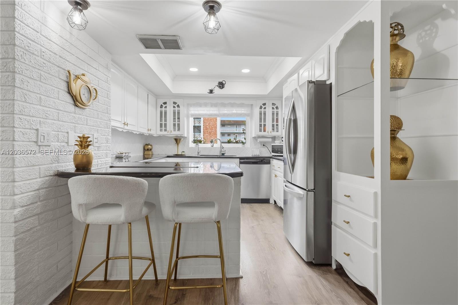 A white kitchen features a breakfast bar with two stools, stainless appliances, and gold accents — 1040 Crystal Lake Dr # 3, Deerfield Beach FL 33064, DEERFIELD BEACH, FL