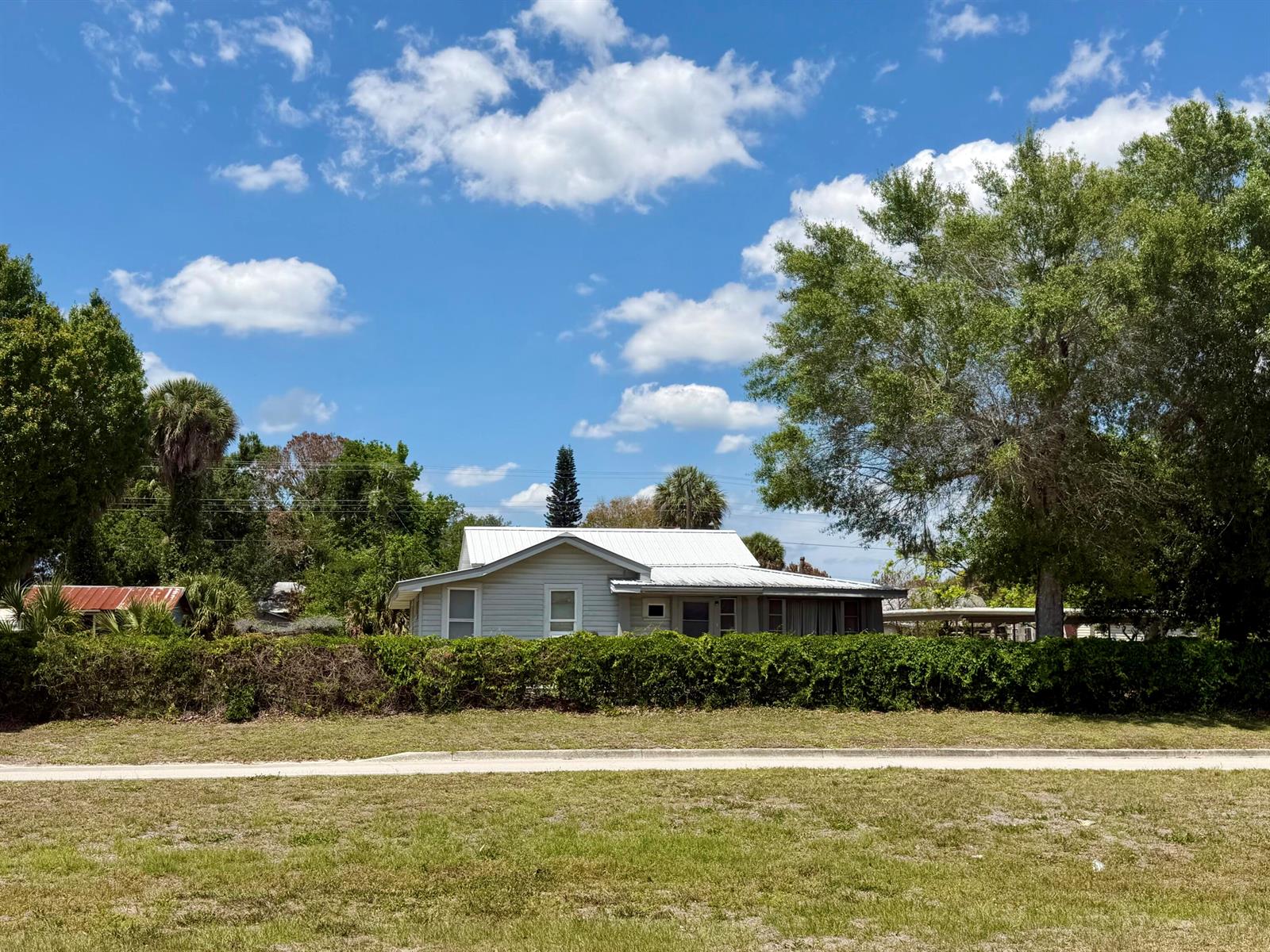 A light-colored house with a metal roof sits behind a tall hedge and trees — 207 SE 4th Street, Okeechobee FL 34974, OKEECHOBEE, FL