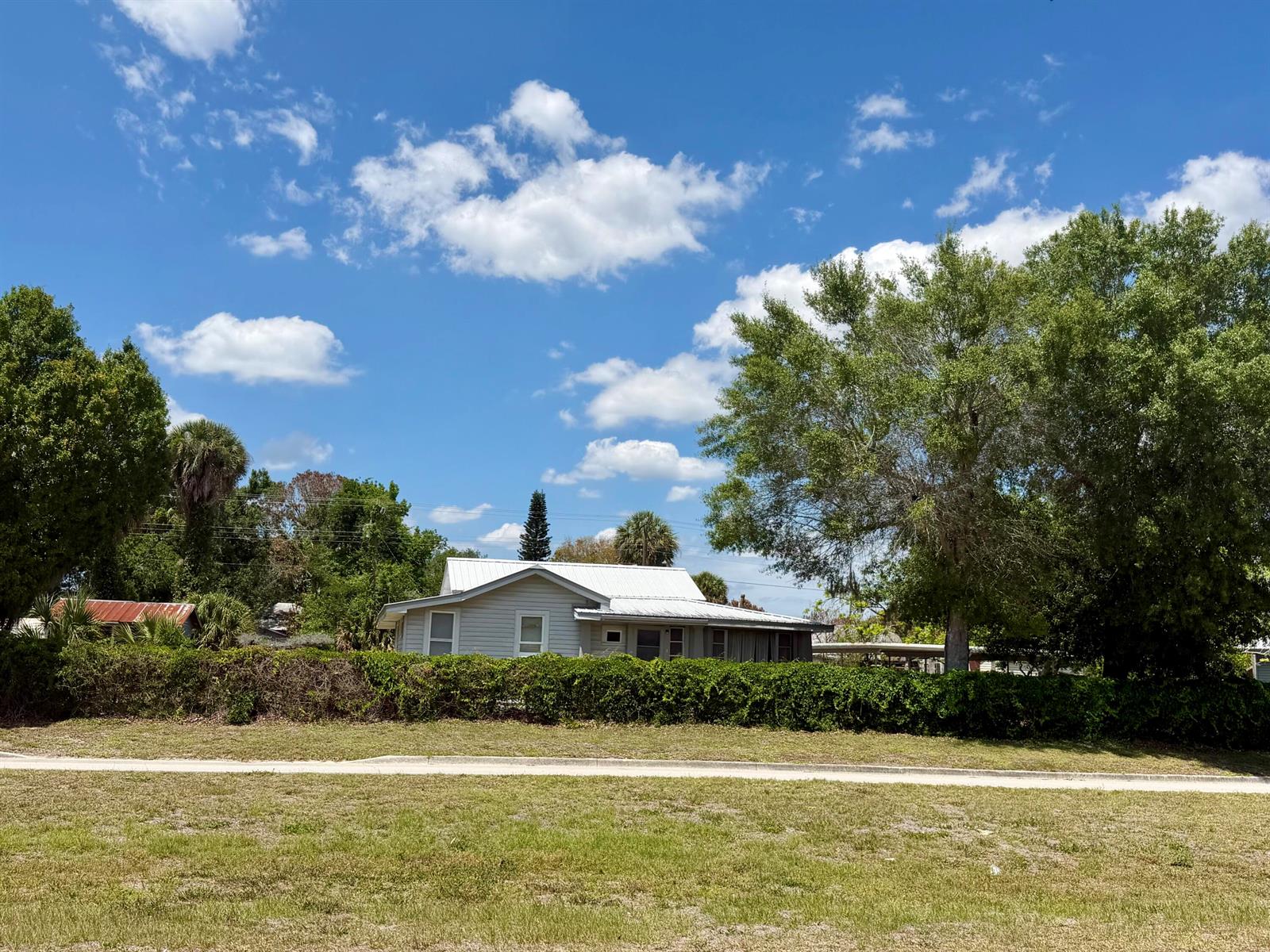 A light-colored house with a white metal roof sits behind a dense green hedge — 207 SE 4th Street, Okeechobee FL 34974, OKEECHOBEE, FL