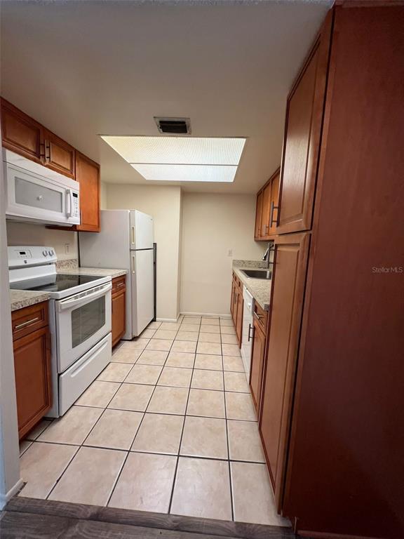 A galley kitchen features wood cabinets, white appliances, and light-colored square floor tiles — 6043 VILLAGE CIRCLE UNIT 6043, ORLANDO, FL
