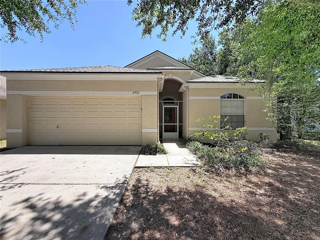 A single-story house with a beige stucco exterior, a two-car garage, and mature trees — 6932 MONARCH PARK DRIVE, APOLLO BEACH, FL