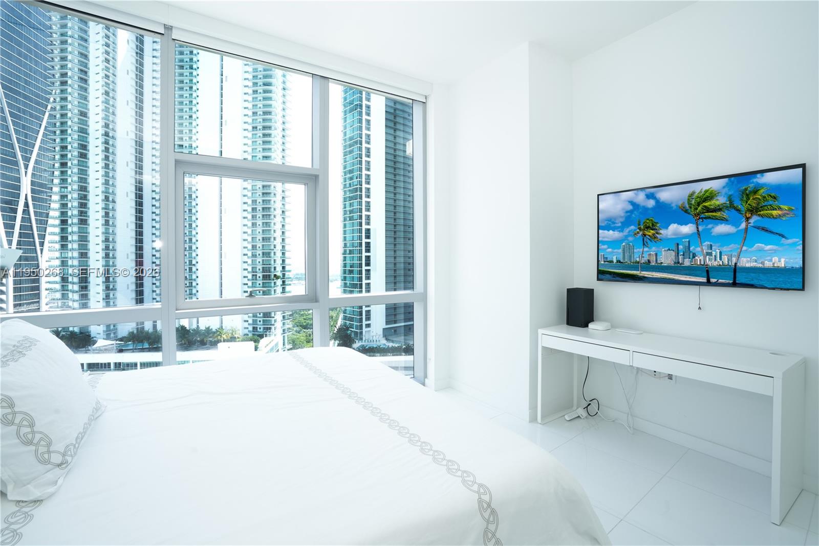 A white bedroom features a wall-mounted television, a desk, and floor-to-ceiling windows overlooking city buildings — 851 NE 1st Ave # 2101, Miami FL 33132, MIAMI, FL