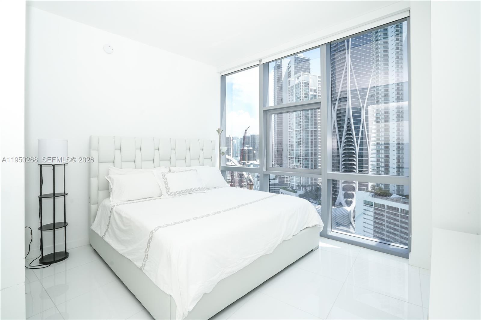 A modern bedroom features a white bed, tiled floors, and floor-to-ceiling windows overlooking city skyscrapers — 851 NE 1st Ave # 2101, Miami FL 33132, MIAMI, FL