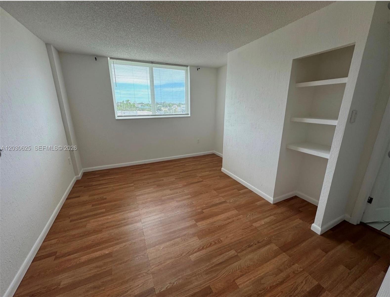 An empty room features wood-look flooring, white walls, a window with blinds, and built-in shelving — 1251 NE 108th St # 409, Miami FL 33161, MIAMI, FL