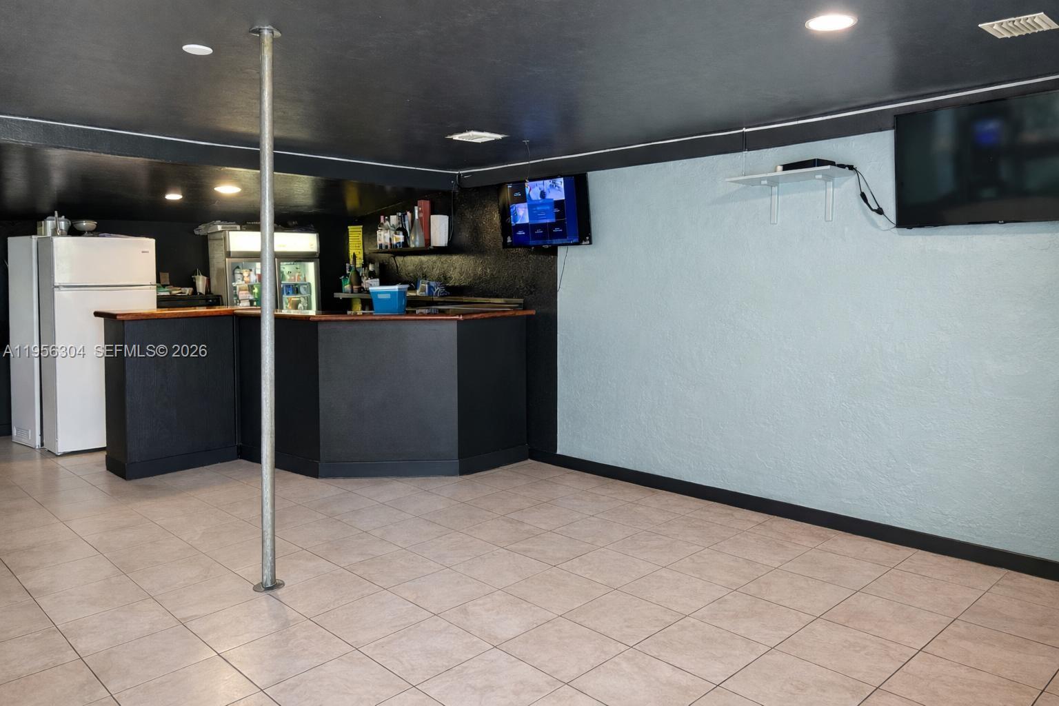 A basement room features a bar, a metal pole, a refrigerator, and a wall-mounted television — 21855 S Dixie Hwy, Miami FL 33170, MIAMI, FL