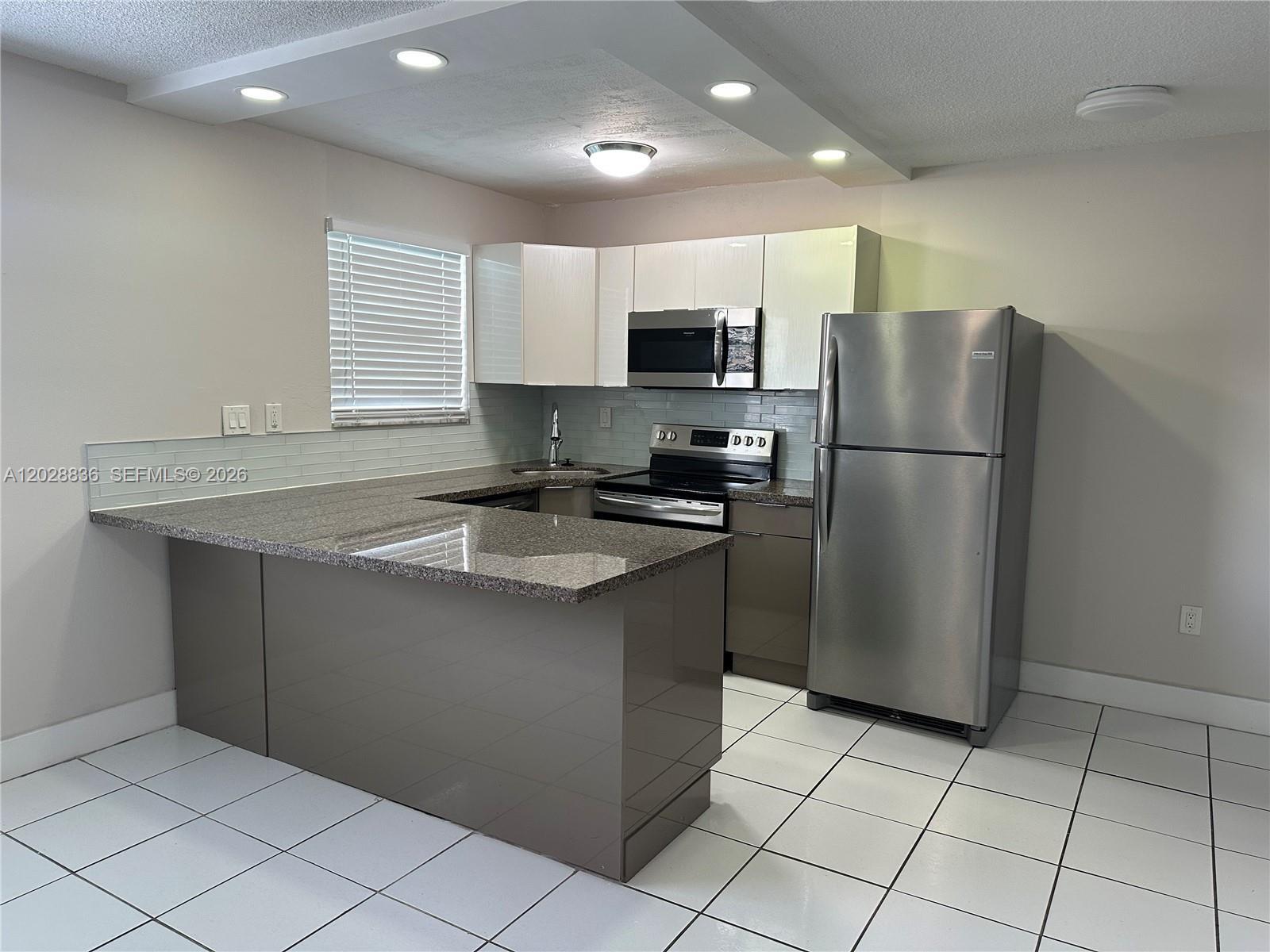 A kitchen features a granite peninsula, stainless steel appliances, and white tile flooring — 1975 NE 135th St # 3E, North Miami FL 33181, NORTH MIAMI, FL
