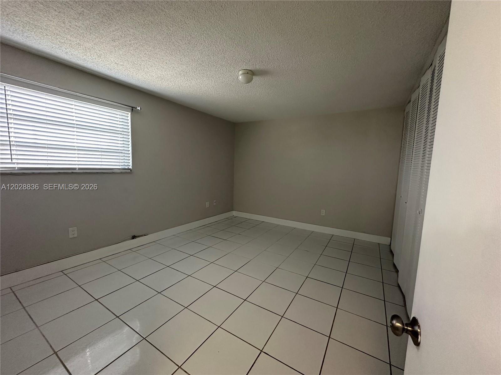 An empty bedroom features light-colored tile flooring, beige walls, a window with blinds, and closet doors — 1975 NE 135th St # 3E, North Miami FL 33181, NORTH MIAMI, FL