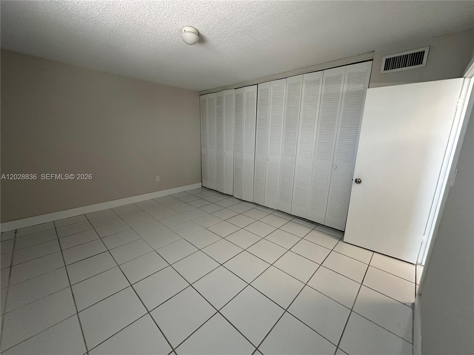 An empty room features white tiled floors, beige walls, and a large white louvered closet — 1975 NE 135th St # 3E, North Miami FL 33181, NORTH MIAMI, FL