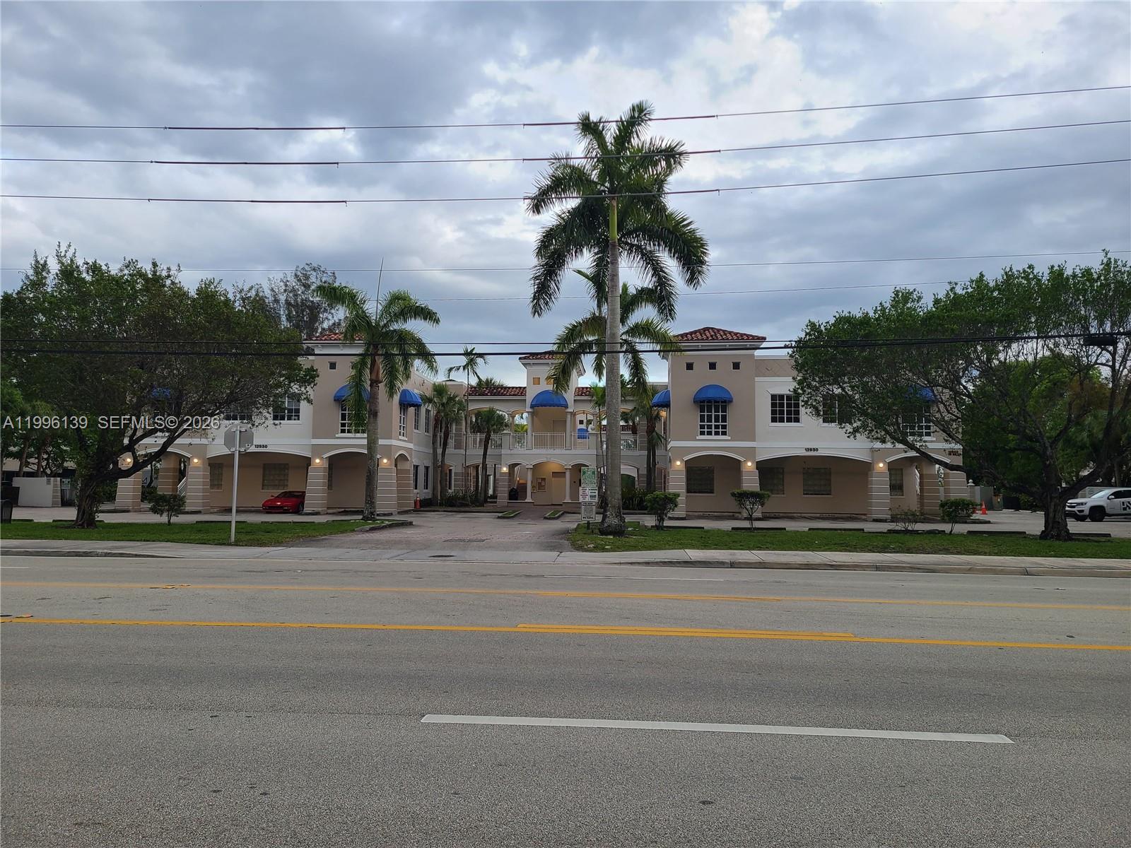 A two-story commercial building with tan stucco walls, blue awnings, and palm trees in front — 12940 SW 128th St # 202, Miami FL 33186, MIAMI, FL