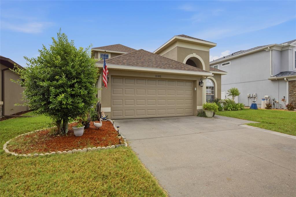 A tan two-story house features a two-car garage, a front lawn, and a small tree — 12452 CRICKLEWOOD DRIVE, SPRING HILL, FL